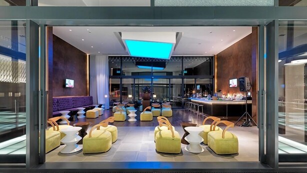 Un moderno vestíbulo de hotel con un bar central, zonas de estar y un gran ventanal.