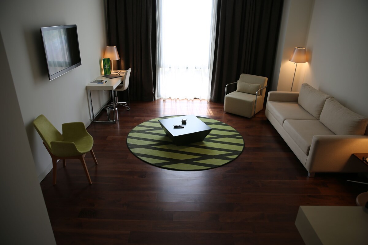 Habitación de hotel moderna con televisión de pantalla plana, un escritorio con lámpara, un sofá, una silla y una mesa de centro.