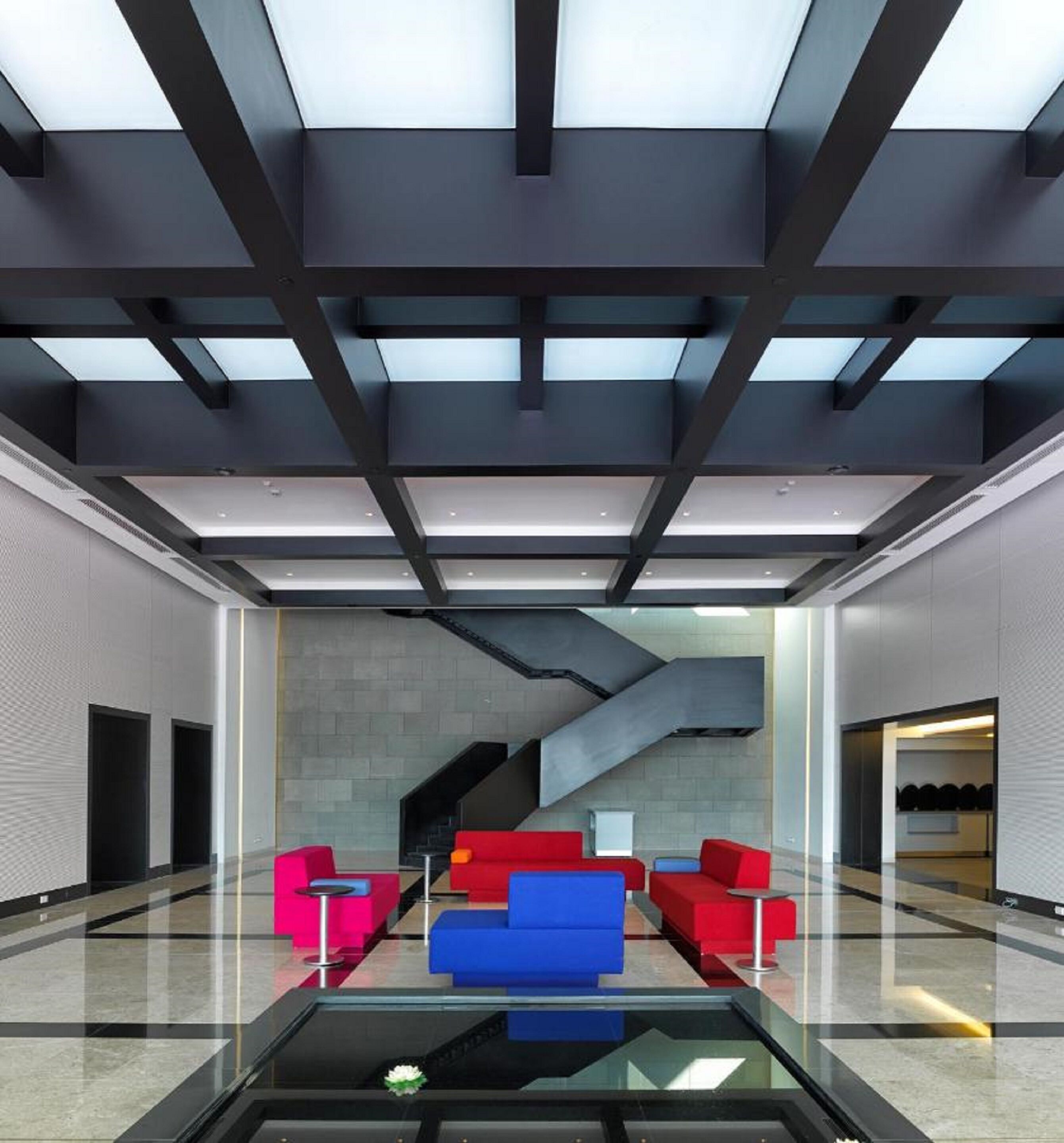 A modern lobby with a glass coffee table, red and blue sofas, a staircase, and a skylight.