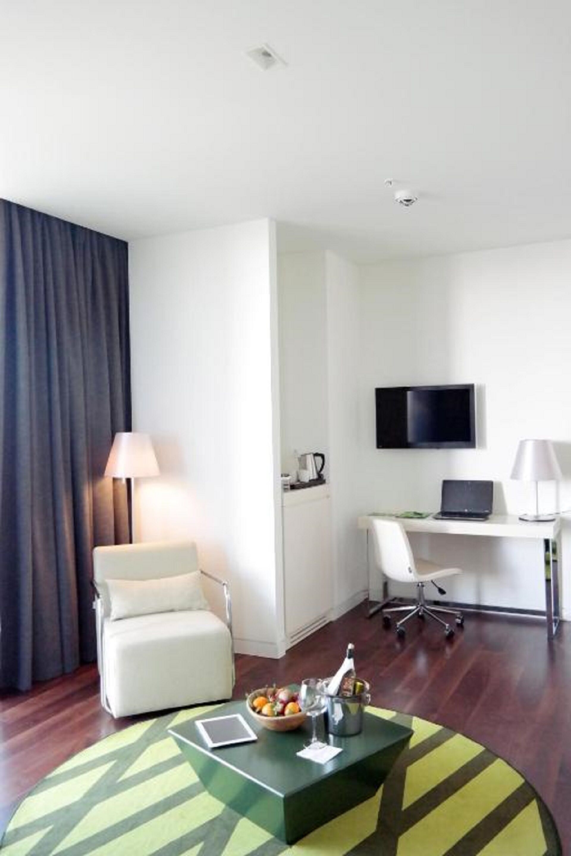 A hotel room with a sofa, a desk with a computer, a TV, and a small table with a bottle and glasses.