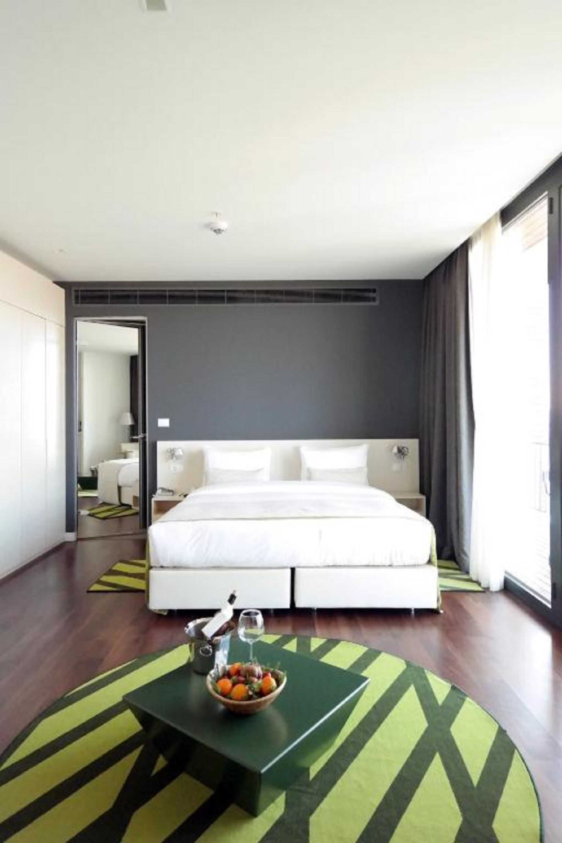 Una habitación de hotel moderna con una cama grande, una mesita de noche con un frutero y una botella, y vistas de la ciudad a través de amplios ventanales.
