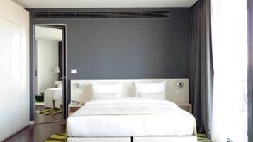 Una habitación de hotel moderna con una cama grande, una mesita de noche con un frutero y una botella, y vistas de la ciudad a través de amplios ventanales.