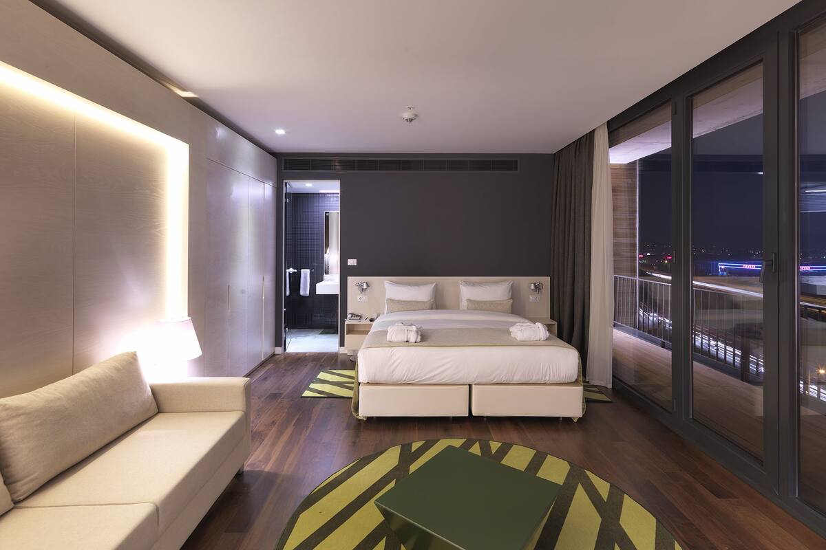 A modern hotel room with a large bed, a sofa, a view of the city at night, and a large window.