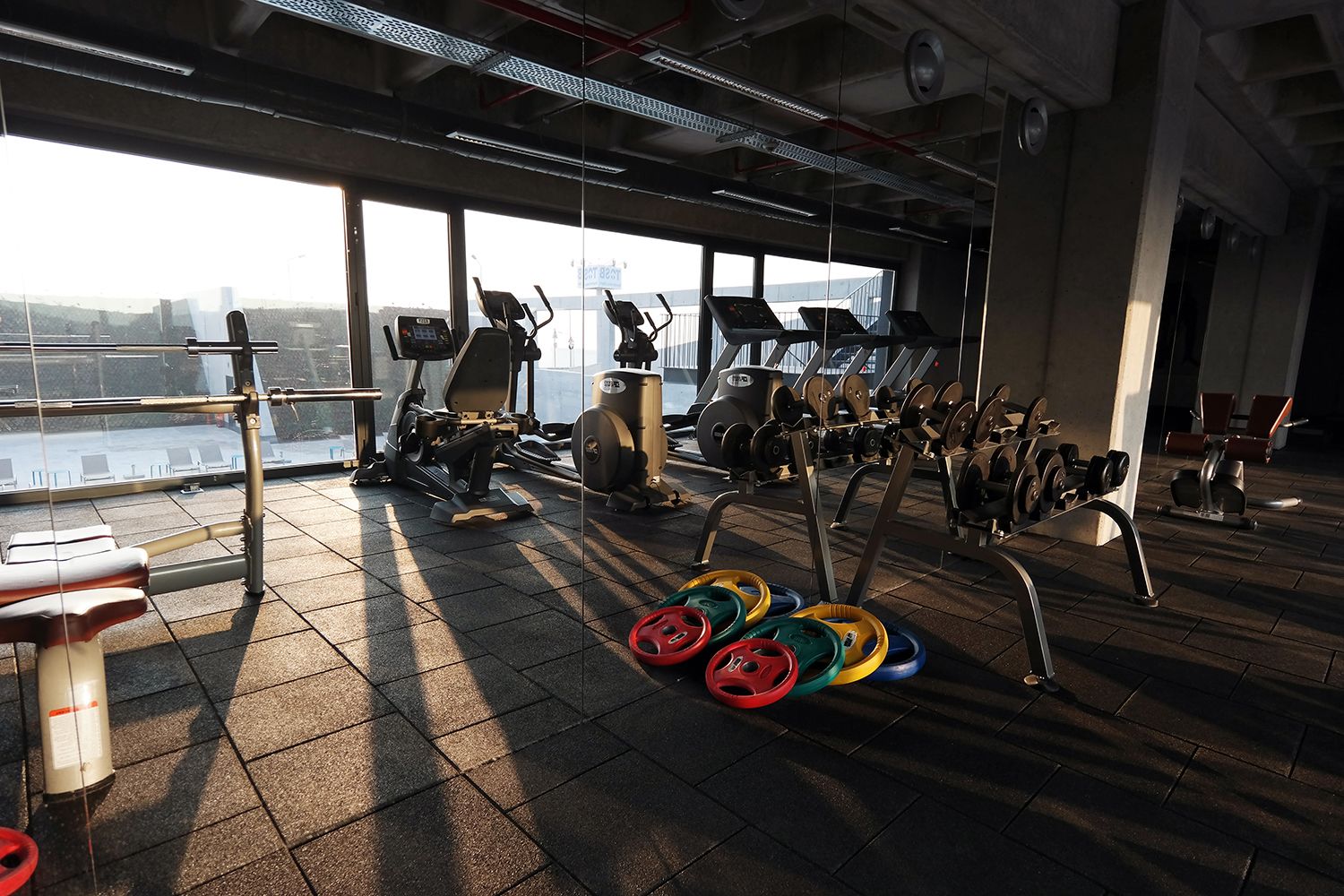 A gym with cardio machines, weightlifting equipment, and a view of a cityscape through large windows.