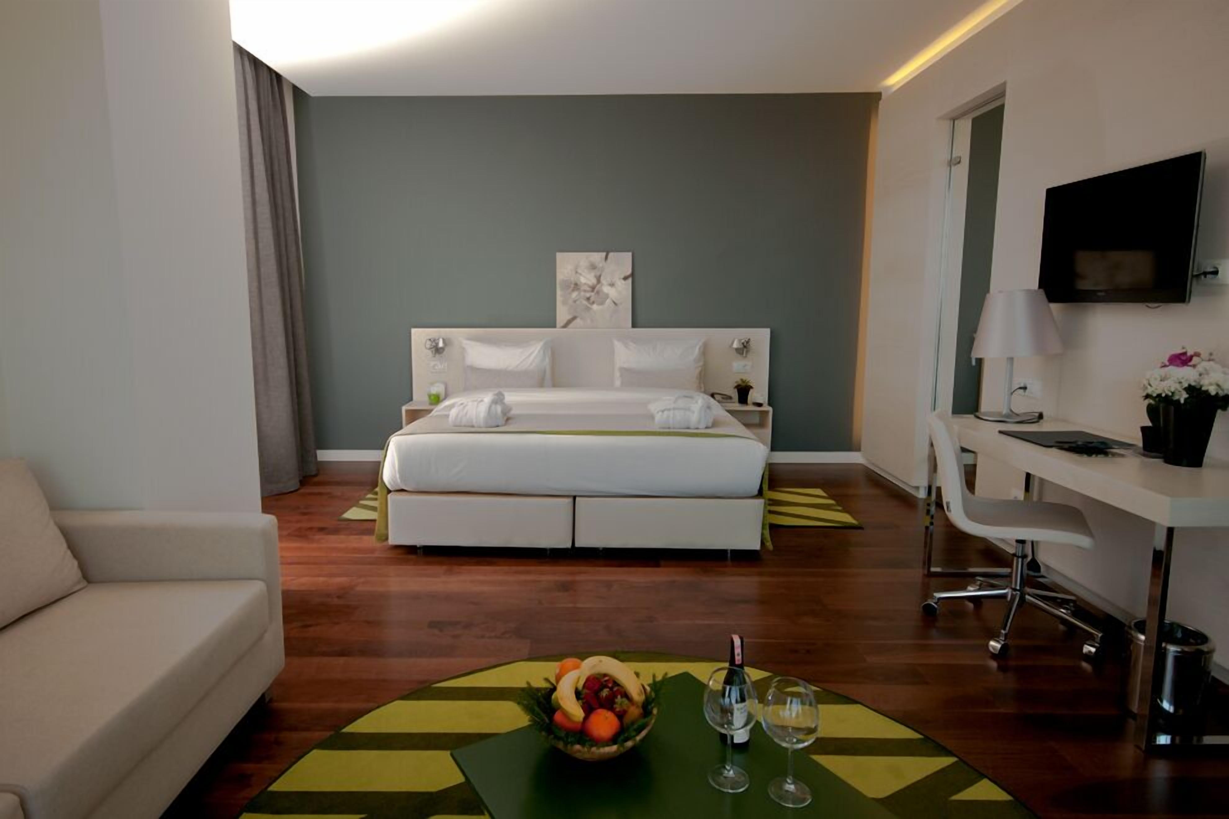 Ein Hotelzimmer mit Bett, Schreibtisch, Sessel, Fernseher und Obstschale mit Weingläsern.