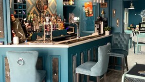 Un área de bar con paredes color turquesa, taburetes, lámparas colgantes y una variedad de botellas y vasos.