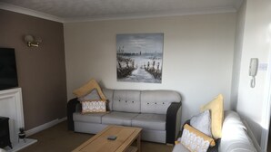 Een woonkamer met een grijze bank, een witte fauteuil, een houten salontafel en een schilderij van een strand.