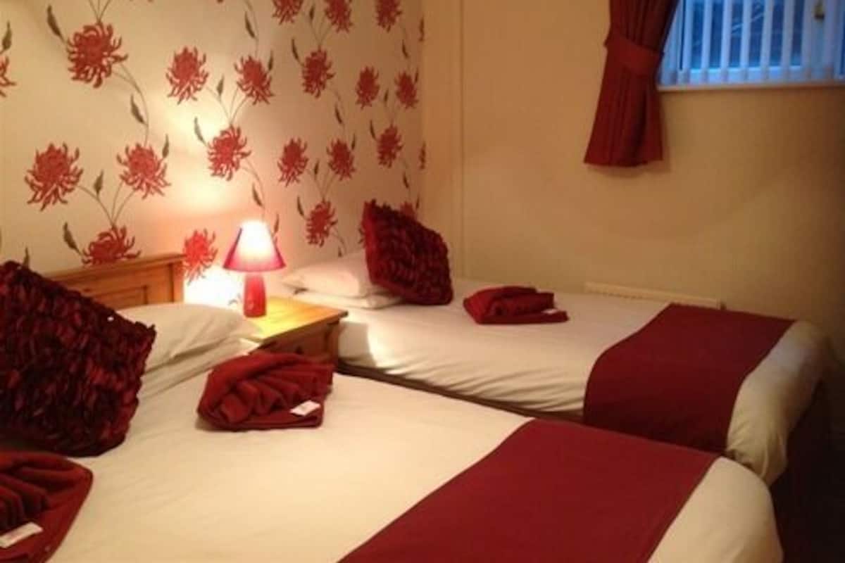 A hotel room with two beds, a nightstand with a lamp, and floral wallpaper.
