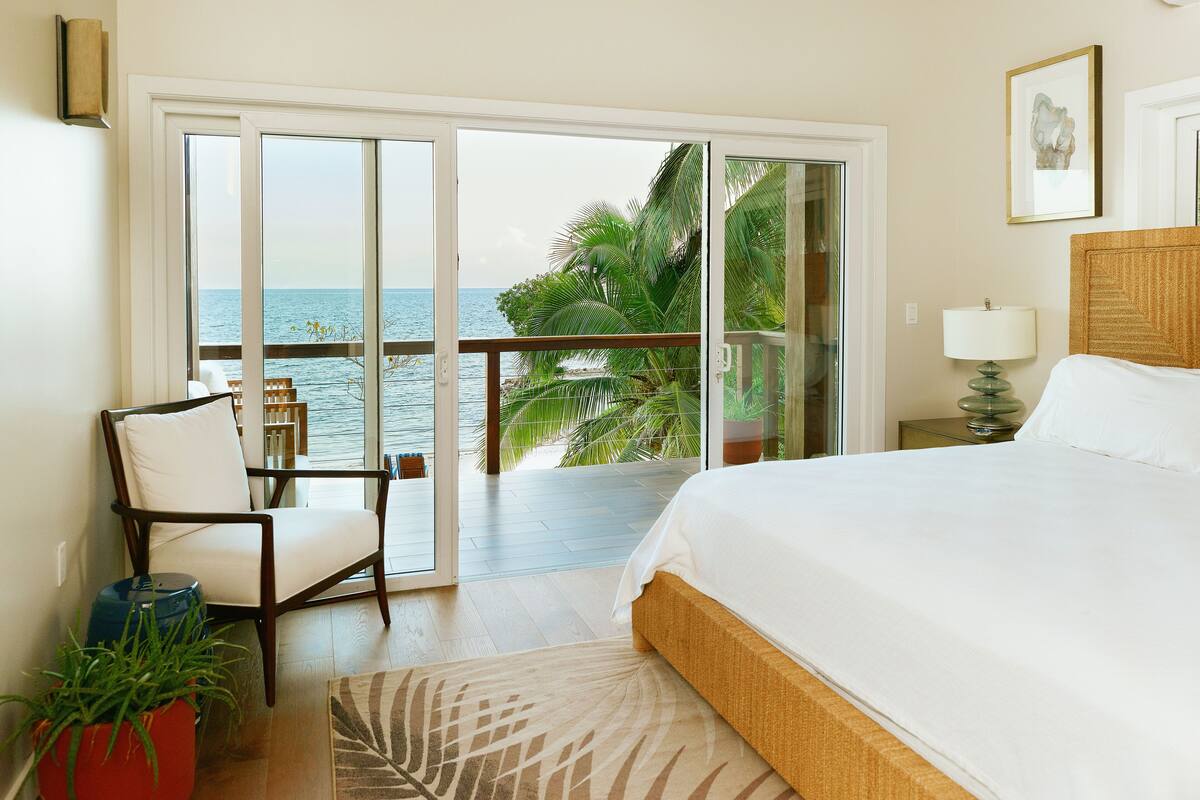 A bedroom with a large bed, a chair, a view of the ocean, and a sliding glass door leading to a balcony.