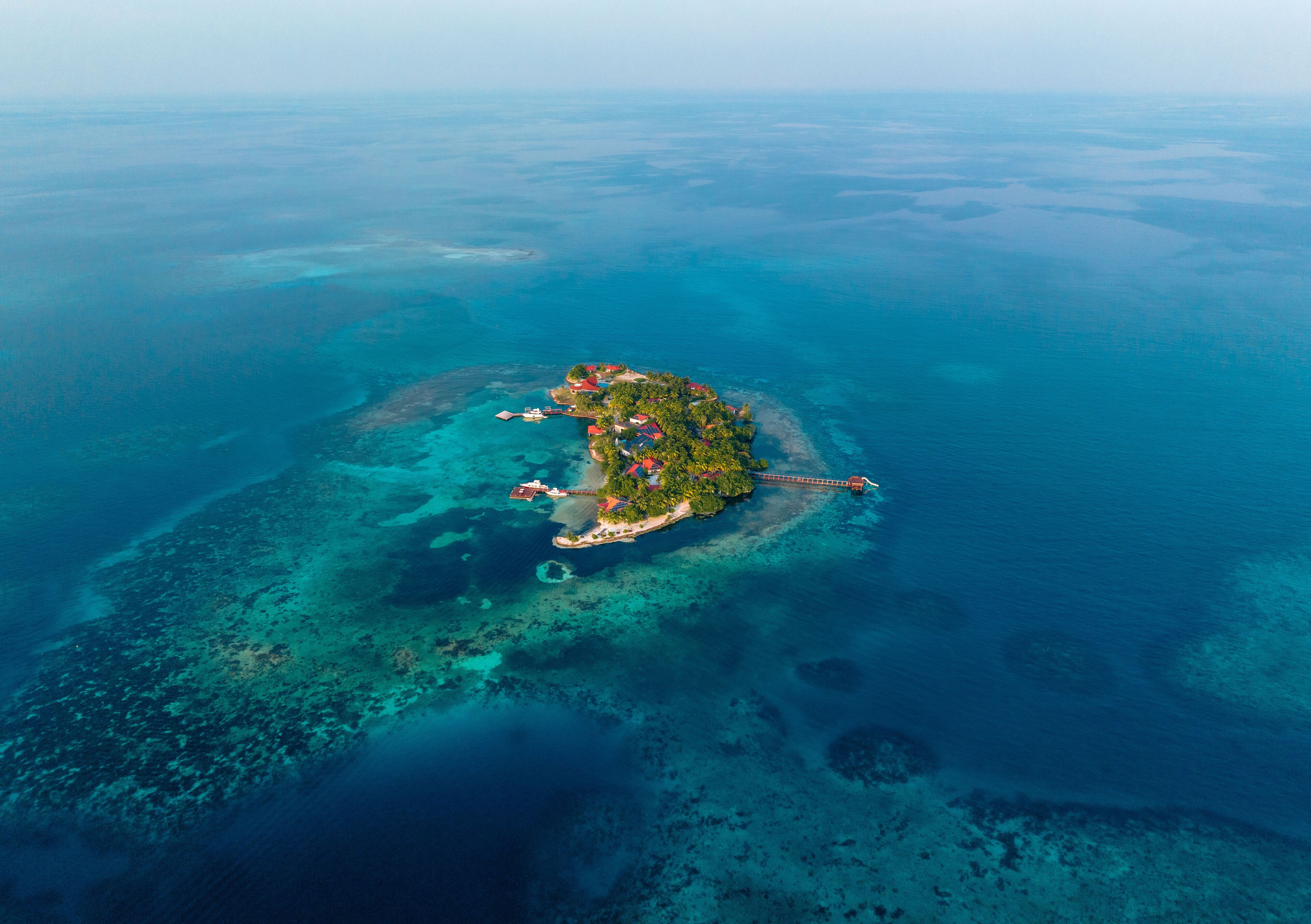 Vue aérienne d’une île tropicale aux eaux d’un bleu limpide, avec une île centrale abritant des bâtiments et une petite péninsule reliée par un pont.