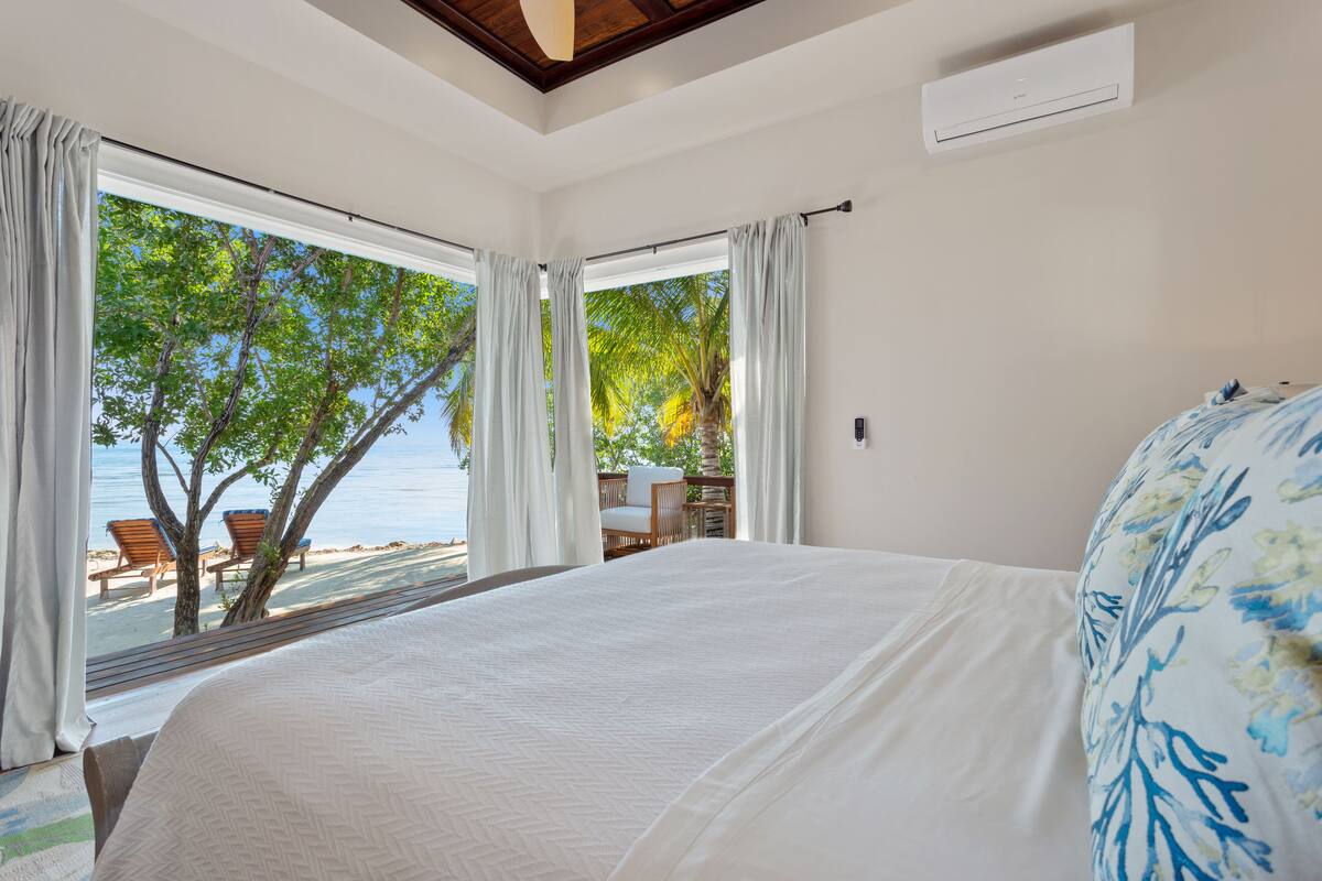 A bedroom with a large bed, white curtains, and a view of the beach and palm trees.