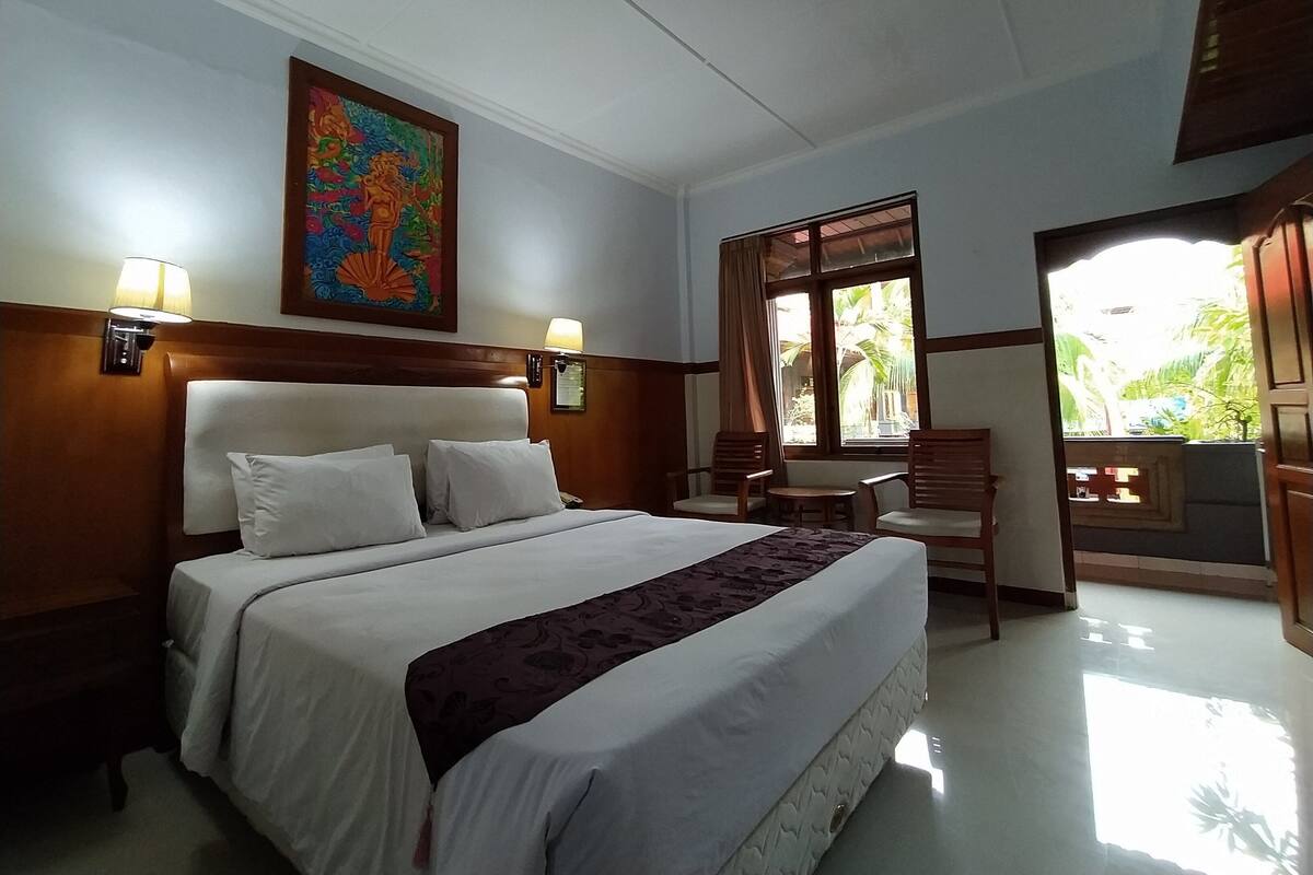A hotel room with a large bed, two bedside lamps, a painting on the wall, a window with curtains, and a small table with chairs.