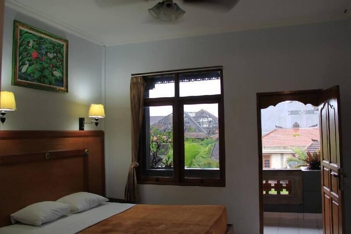 A bedroom with a bed, two bedside lamps, a ceiling fan, a window with a view of buildings, and a door leading to an outdoor area.