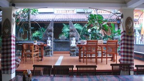 A restaurant with a traditional design, featuring wooden furniture and a tiled floor. There are several tables and chairs arranged for dining, and the space is decorated with plants and statues. - Hotel Sorga (Kuta)
