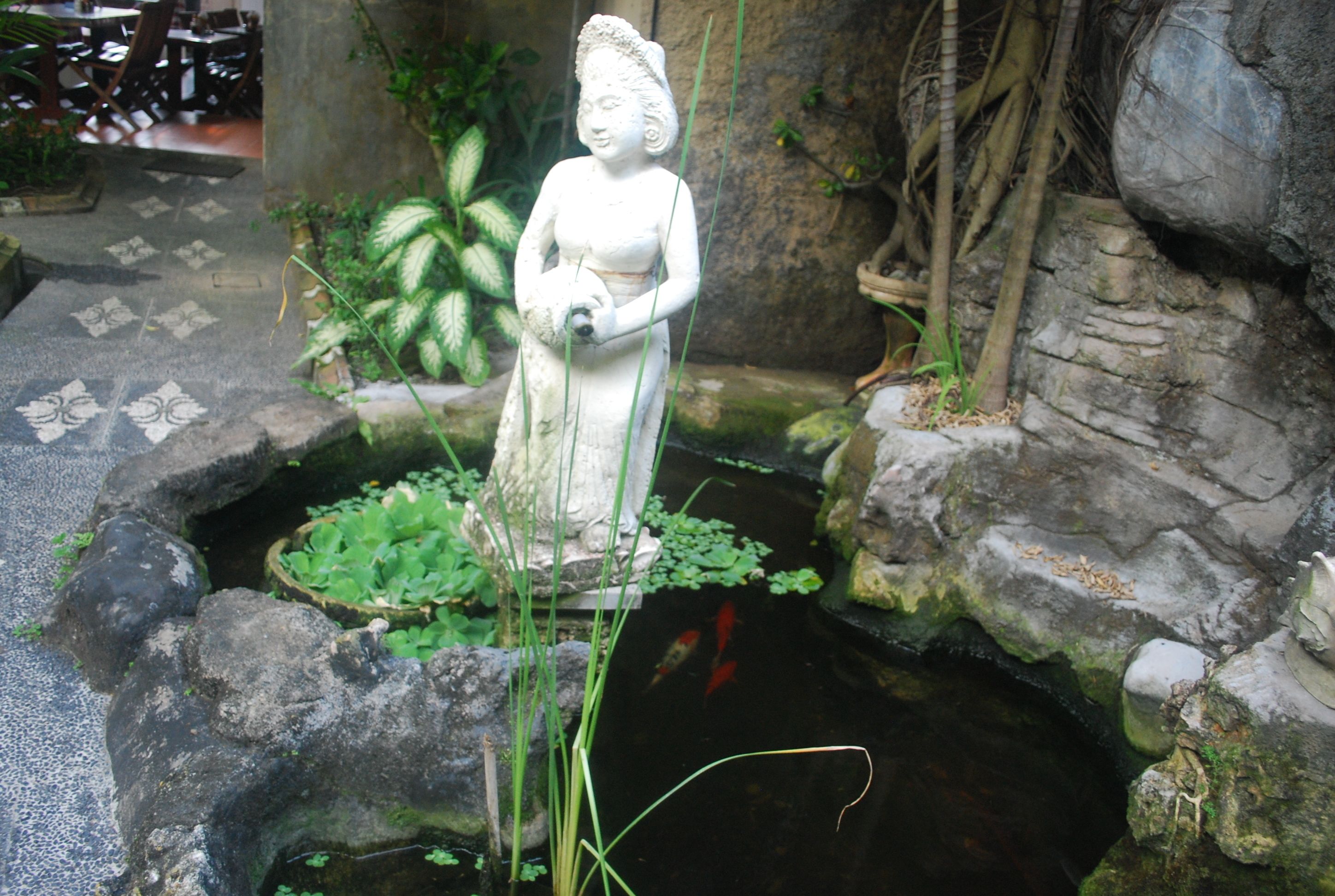A stone statue in a garden with a pond, surrounded by plants and rocks.