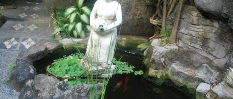 A stone statue in a garden with a pond, surrounded by plants and rocks.
