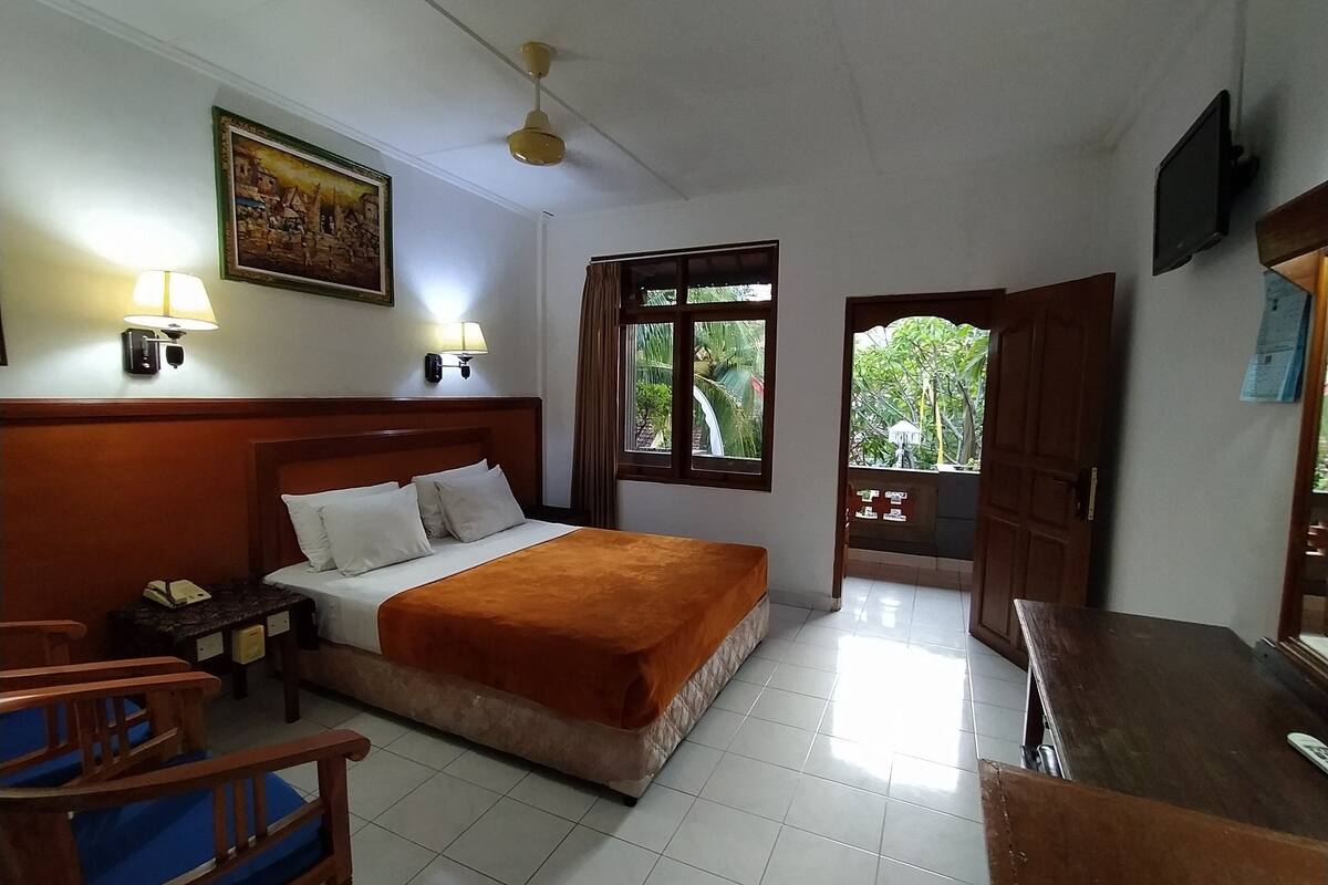 A hotel room with a bed, bedside lamps, a desk, and a view of greenery outside.