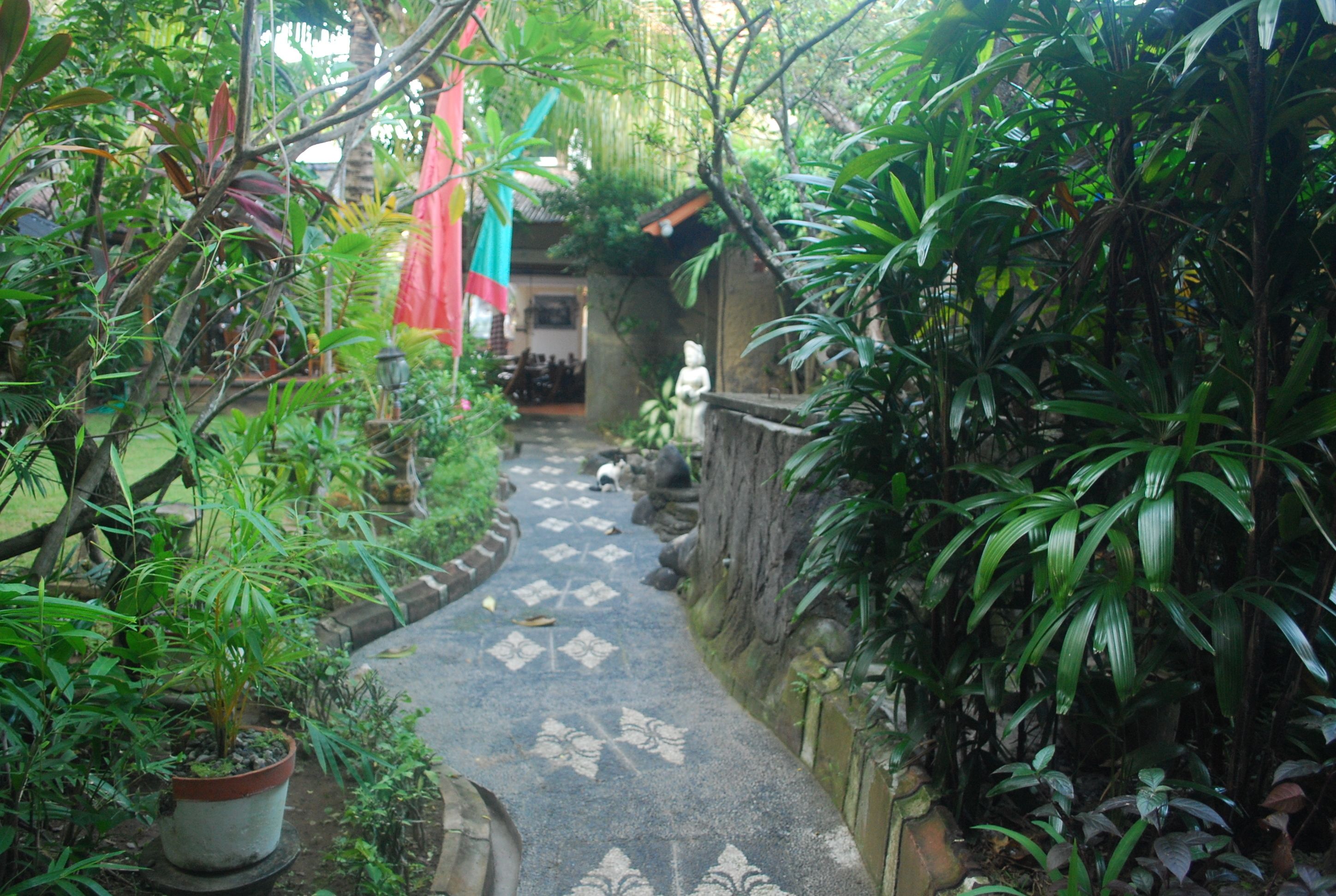 A narrow pathway flanked by lush greenery and a patterned tile floor leading to a building with a tiled roof.