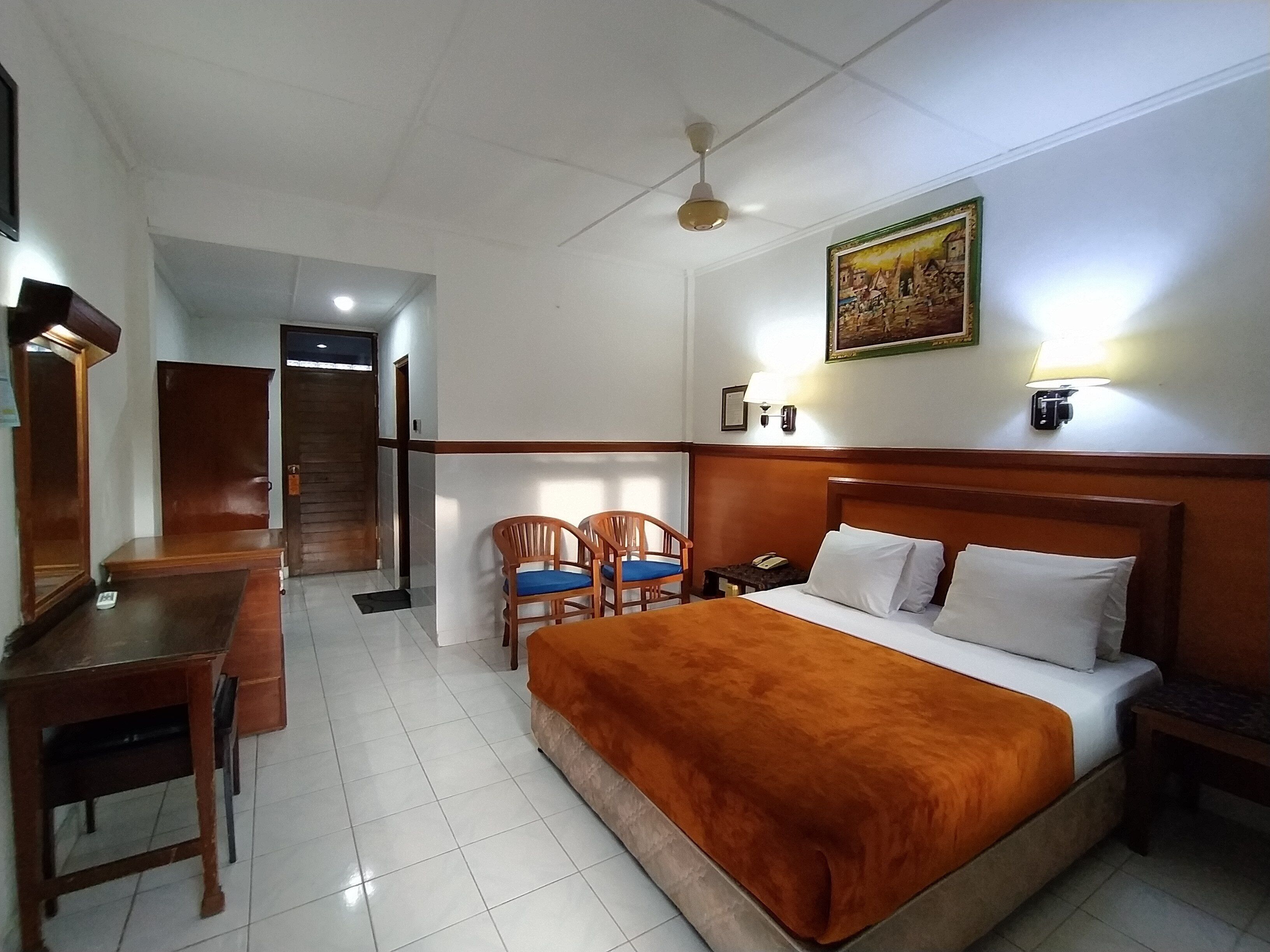 A hotel room with a bed, a desk, two chairs, a nightstand, a lamp, a ceiling fan, and a painting on the wall.