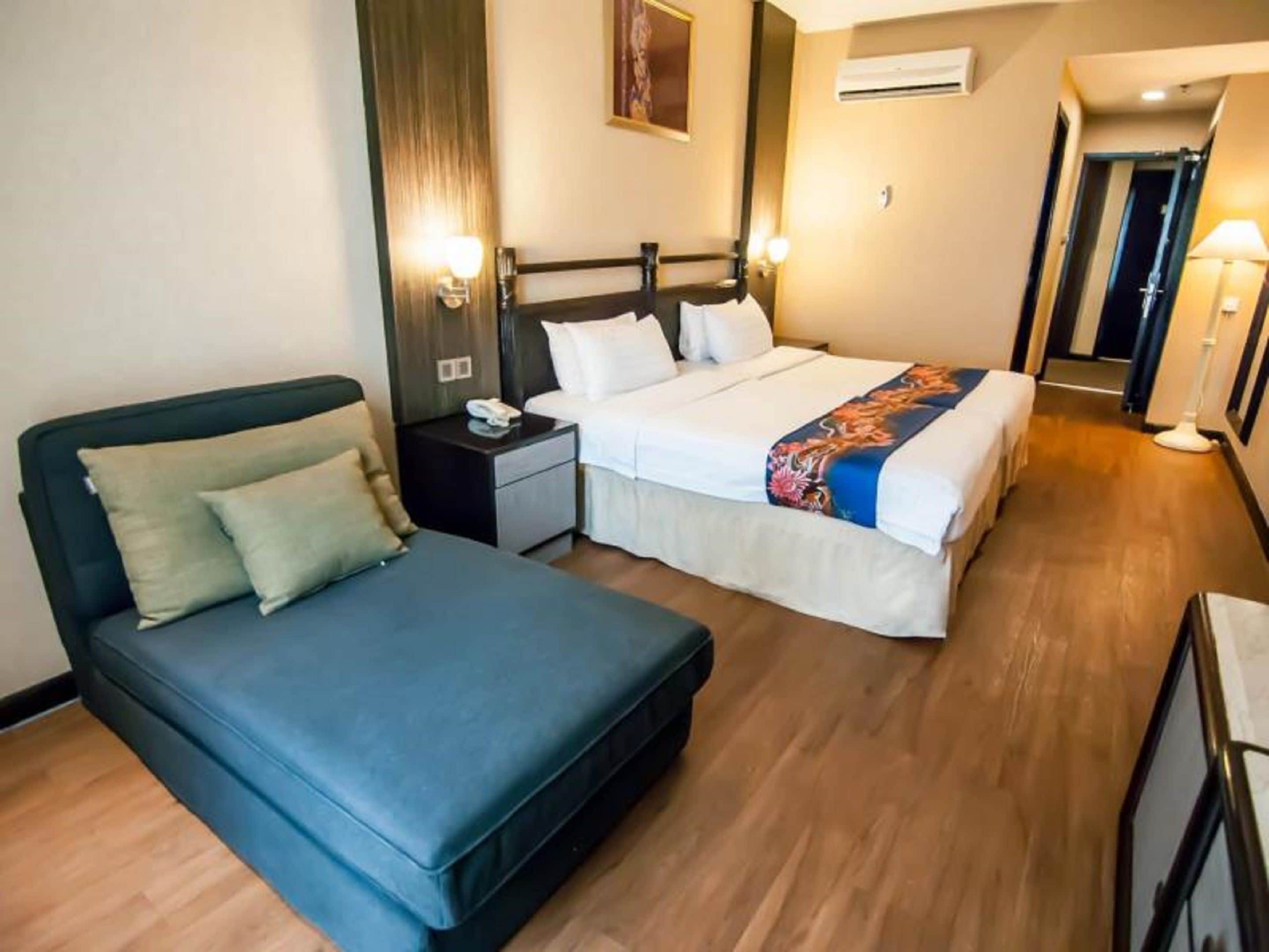 A hotel room with a single bed, a sofa bed, a bedside table with a lamp, and a wall-mounted air conditioner.