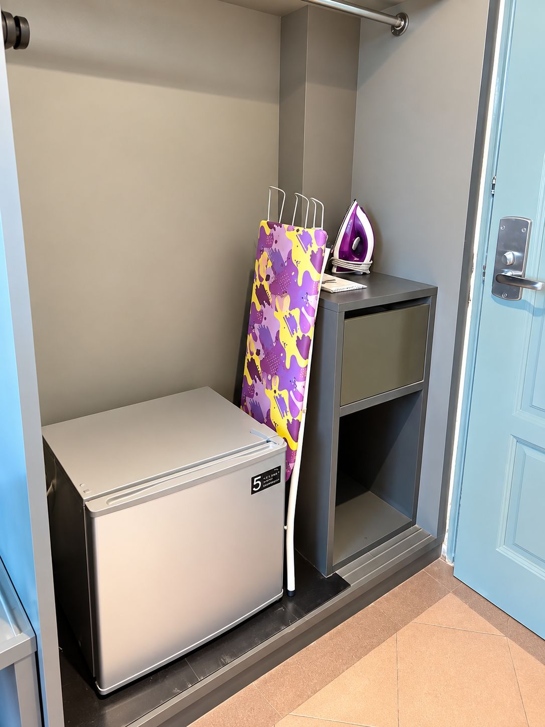 Una kitchenette moderna y compacta con refrigerador integrado, un pequeño centro de planchado y una puerta con manija.