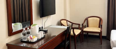 A hotel room with a wooden desk, a television, two chairs, a white trash bin, and an air conditioning unit.