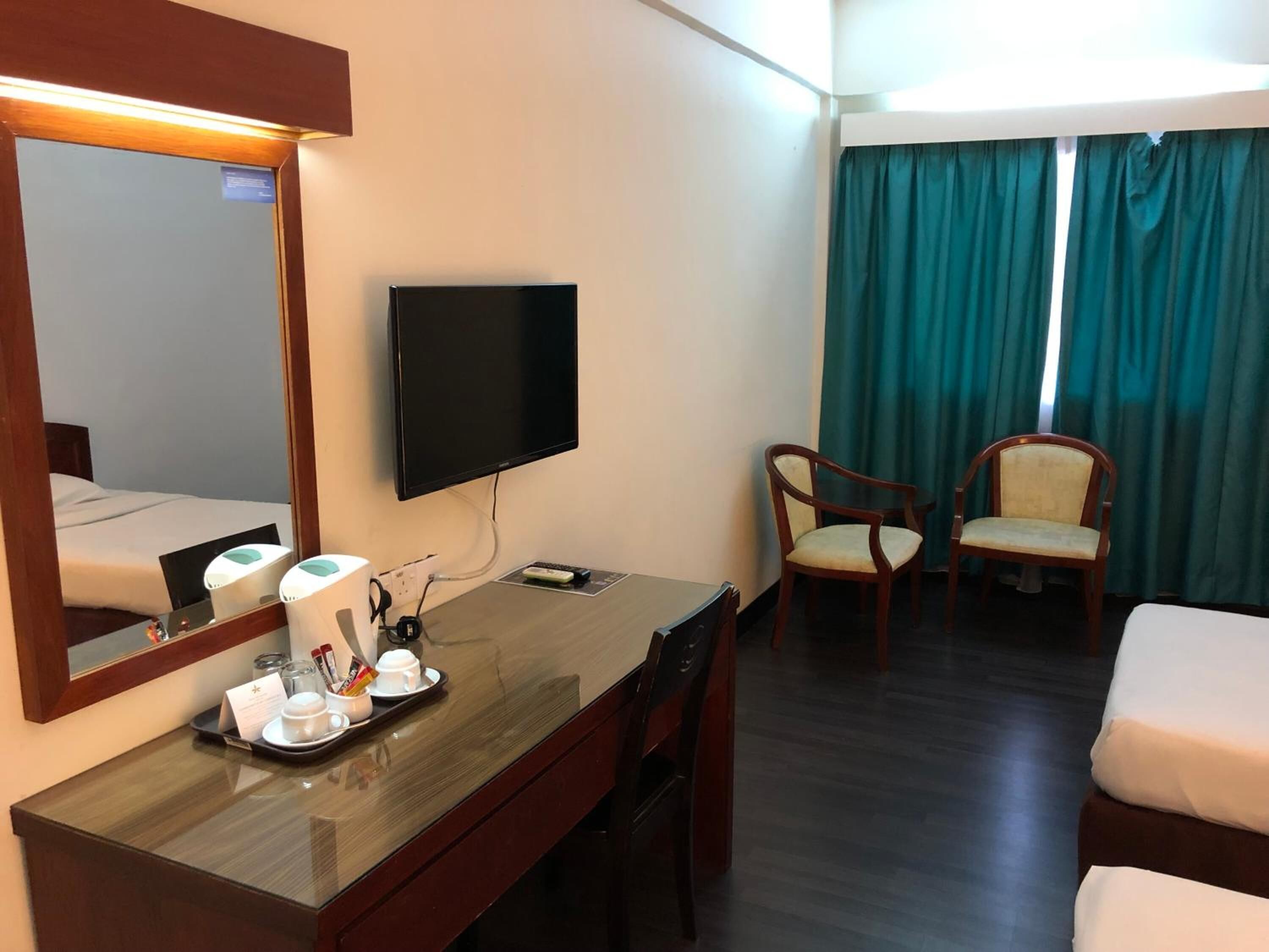 Quarto de hotel com cama, escrivaninha com espelho, televisão, duas cadeiras e janela com cortinas.