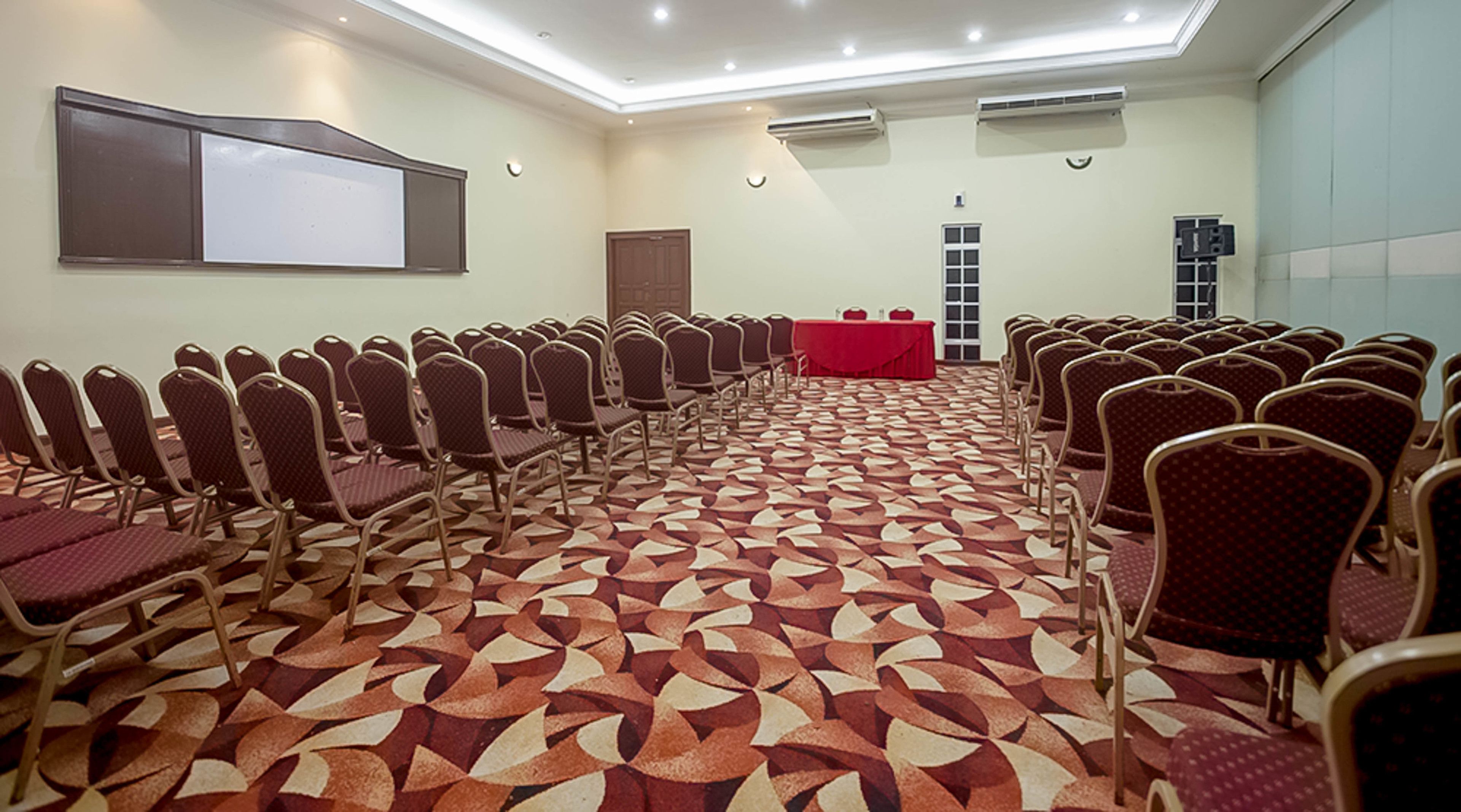 A conference room with rows of chairs, a red table, and a projector screen on the wall.