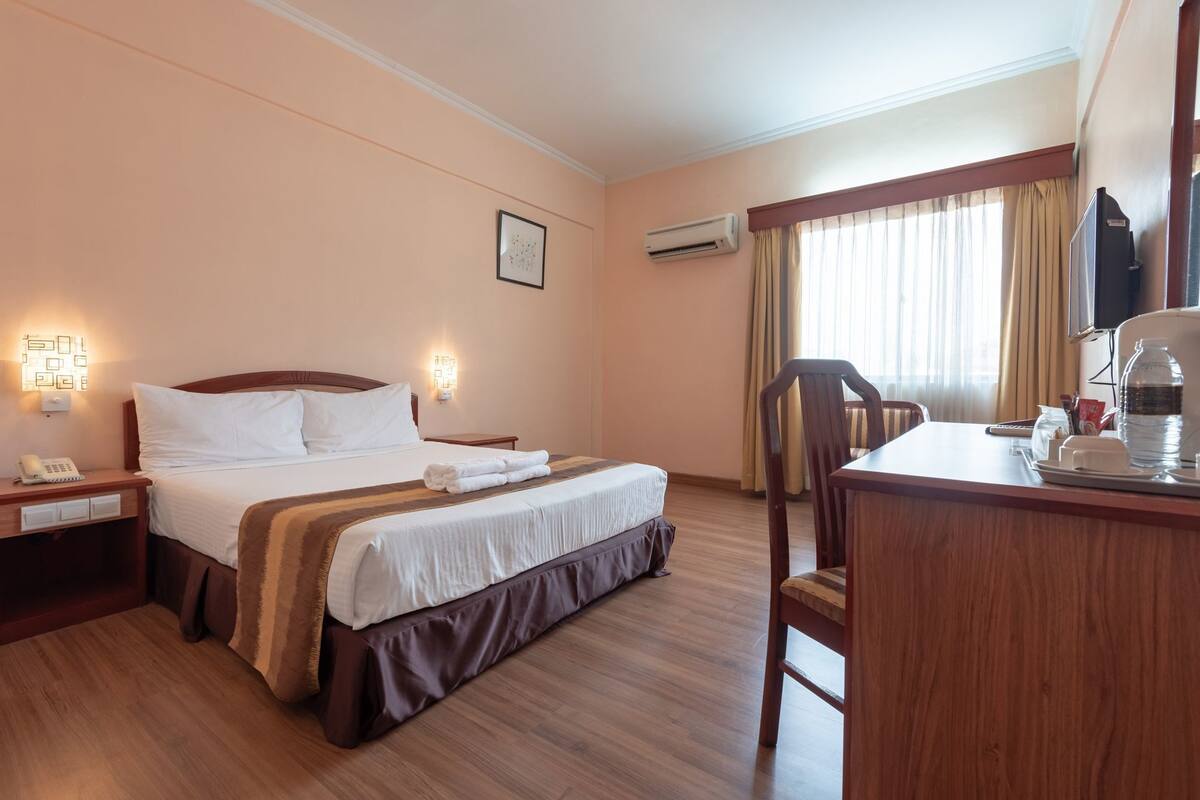 A hotel room with a bed, bedside tables, a desk with a chair, an air conditioning unit, and a window with curtains.