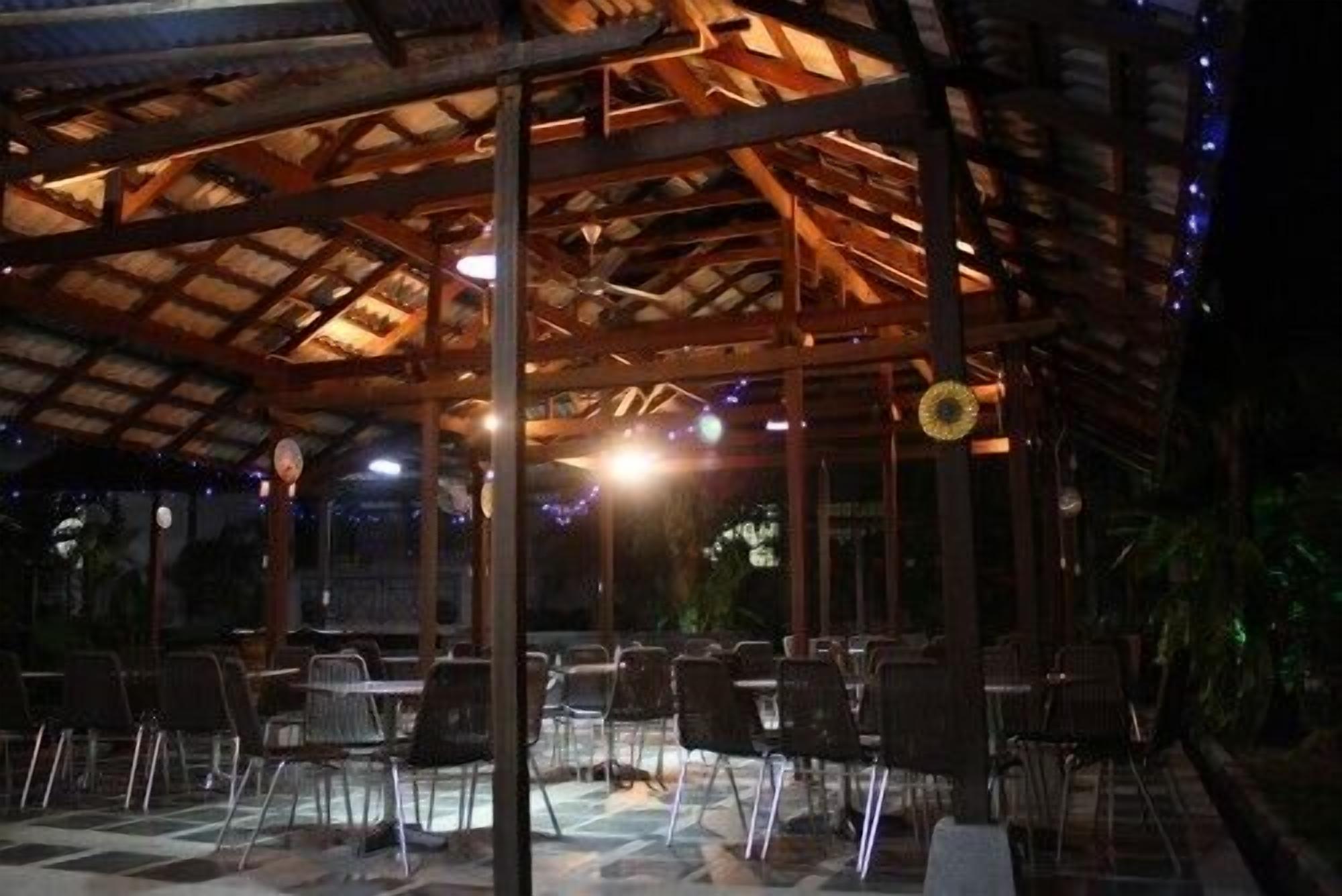 Un patio techado al aire libre con mesas y sillas, vigas de madera y luces colgantes.