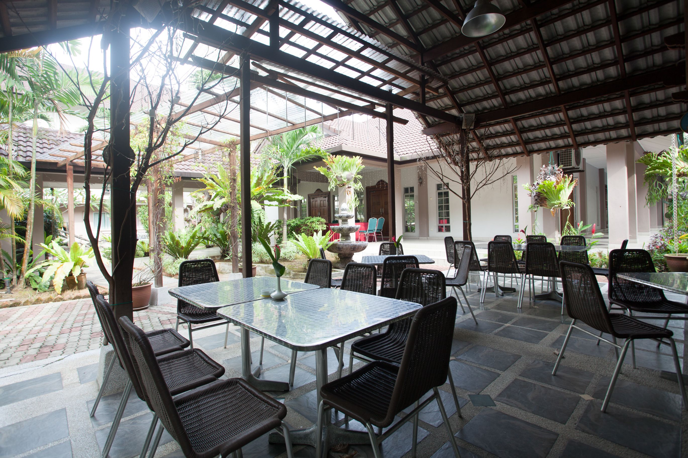 An outdoor patio with a covered seating area, including tables and chairs, surrounded by potted plants and a view of a traditional building in the background.