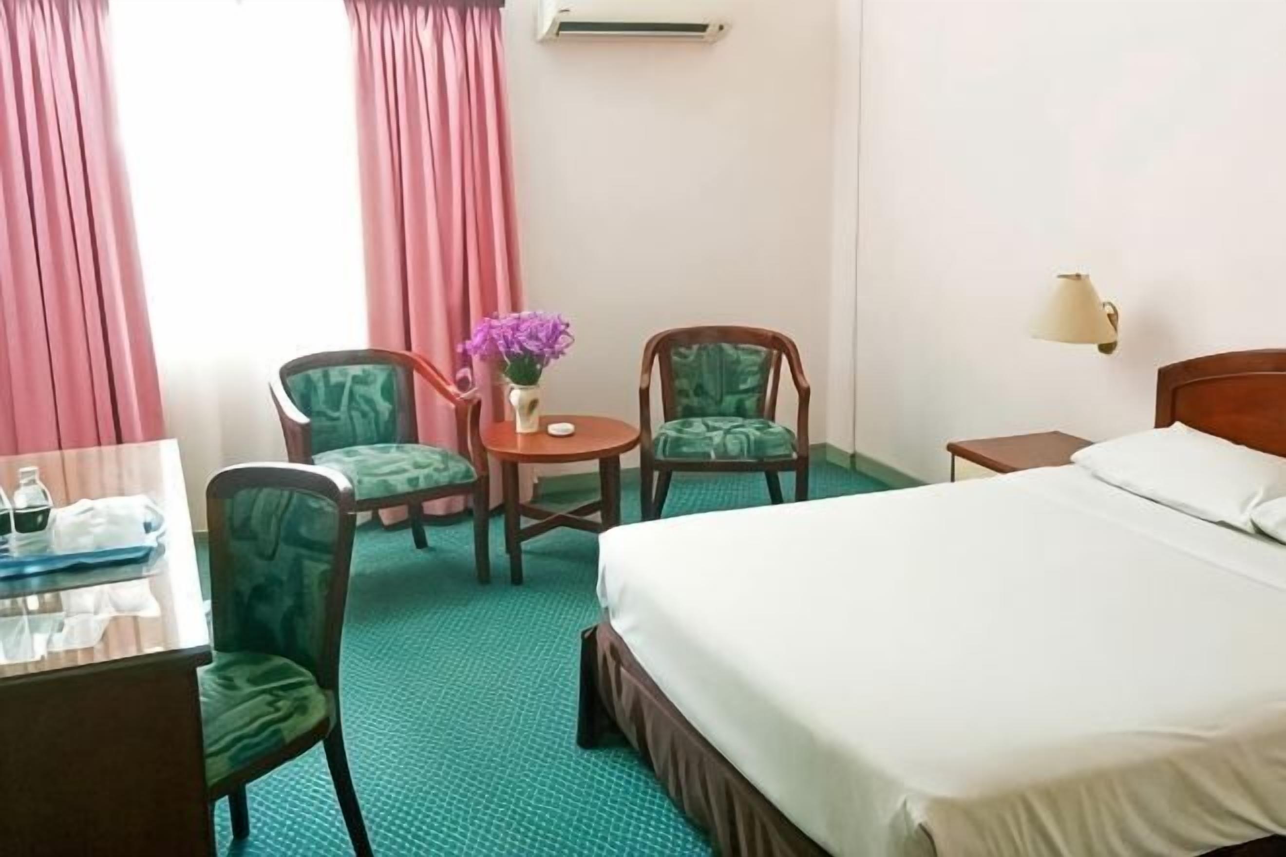 Ett hotellrum med en säng, två gröna stoppade fåtöljer, ett litet runt bord med en vas med blommor, ett skrivbord med ett bricka och ett nattduksbord med en lampa.