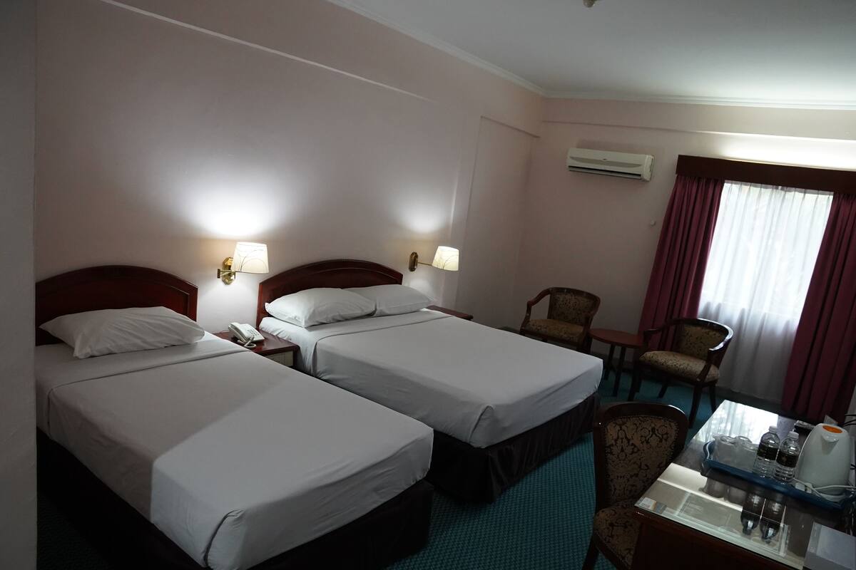A hotel room with two single beds, a desk with a chair, a table with a tray of bottles, and a window with curtains.