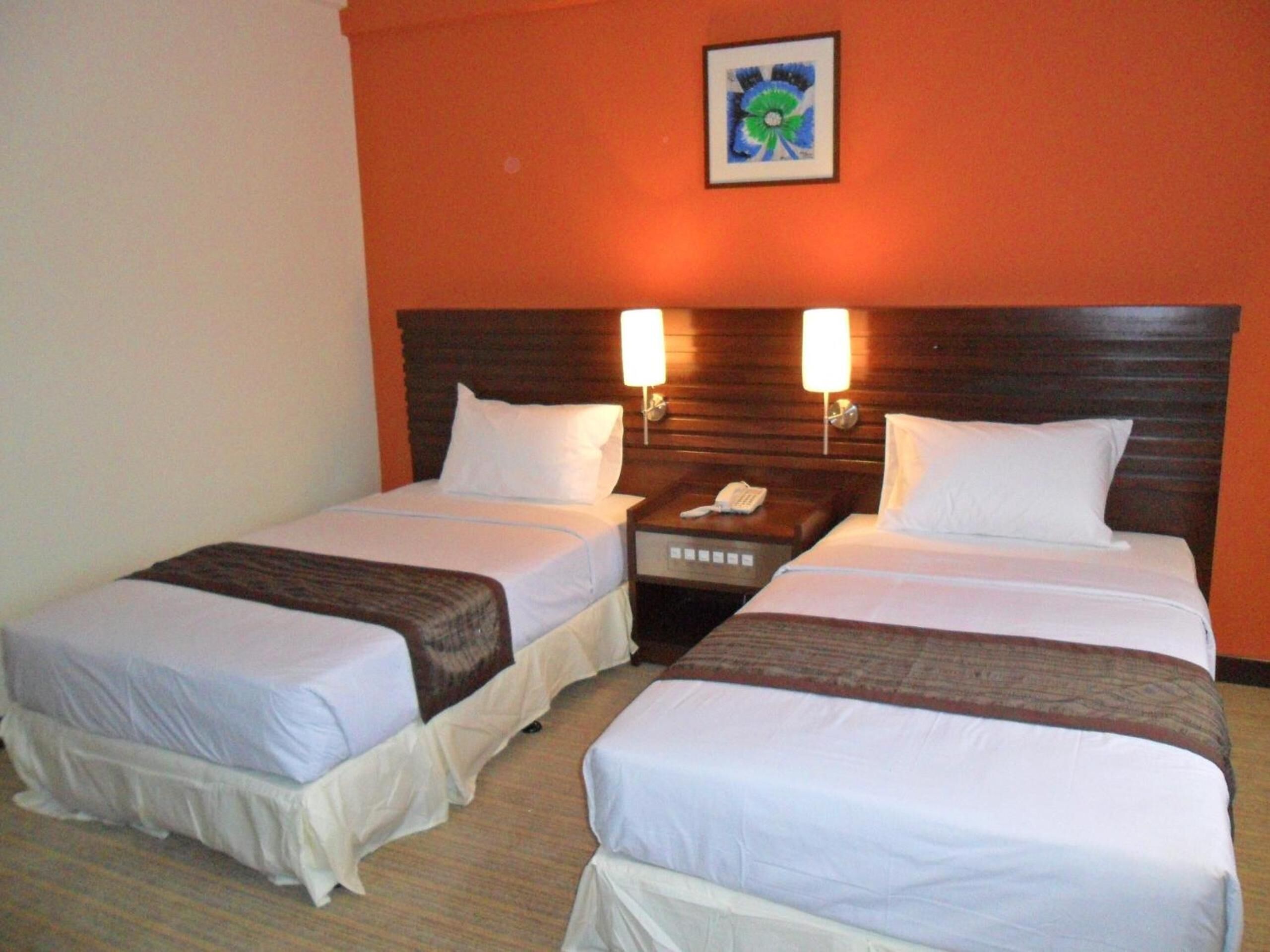 A hotel room with two single beds, each with a white bedspread and a brown headboard. There are two wall-mounted nightstands with lamps, and a small table between the beds. A framed picture hangs on the wall above the beds.
