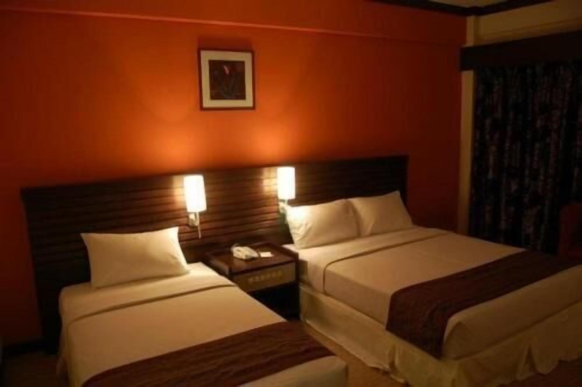 A hotel room with two beds, each with white linens and a brown throw blanket. There is a nightstand with a phone and a lamp between the beds. The room has an orange wall and a framed picture above the beds.