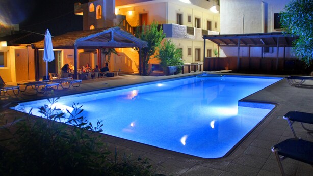 An outdoor swimming pool with lounge chairs and umbrellas, surrounded by buildings with multiple floors and balconies. - Eleonora Anissaras (Hersonissos)