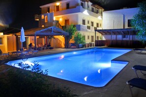 An outdoor swimming pool with lounge chairs and umbrellas, surrounded by buildings with multiple floors and balconies. - Eleonora Anissaras (Hersonissos)