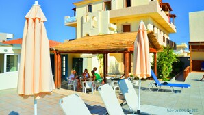 A hotel with outdoor seating and lounge chairs, a covered patio area, and a poolside setting. - Eleonora Anissaras (Hersonissos)