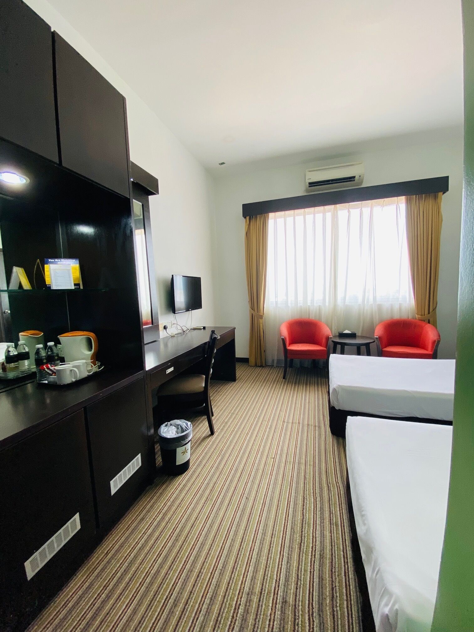 A hotel room with a bed, a desk with a chair, a TV, and two red chairs.