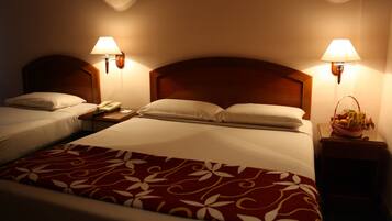 A hotel room with two beds, each with a wooden headboard, bedside lamps, and a telephone. A fruit basket is visible on the nightstand.