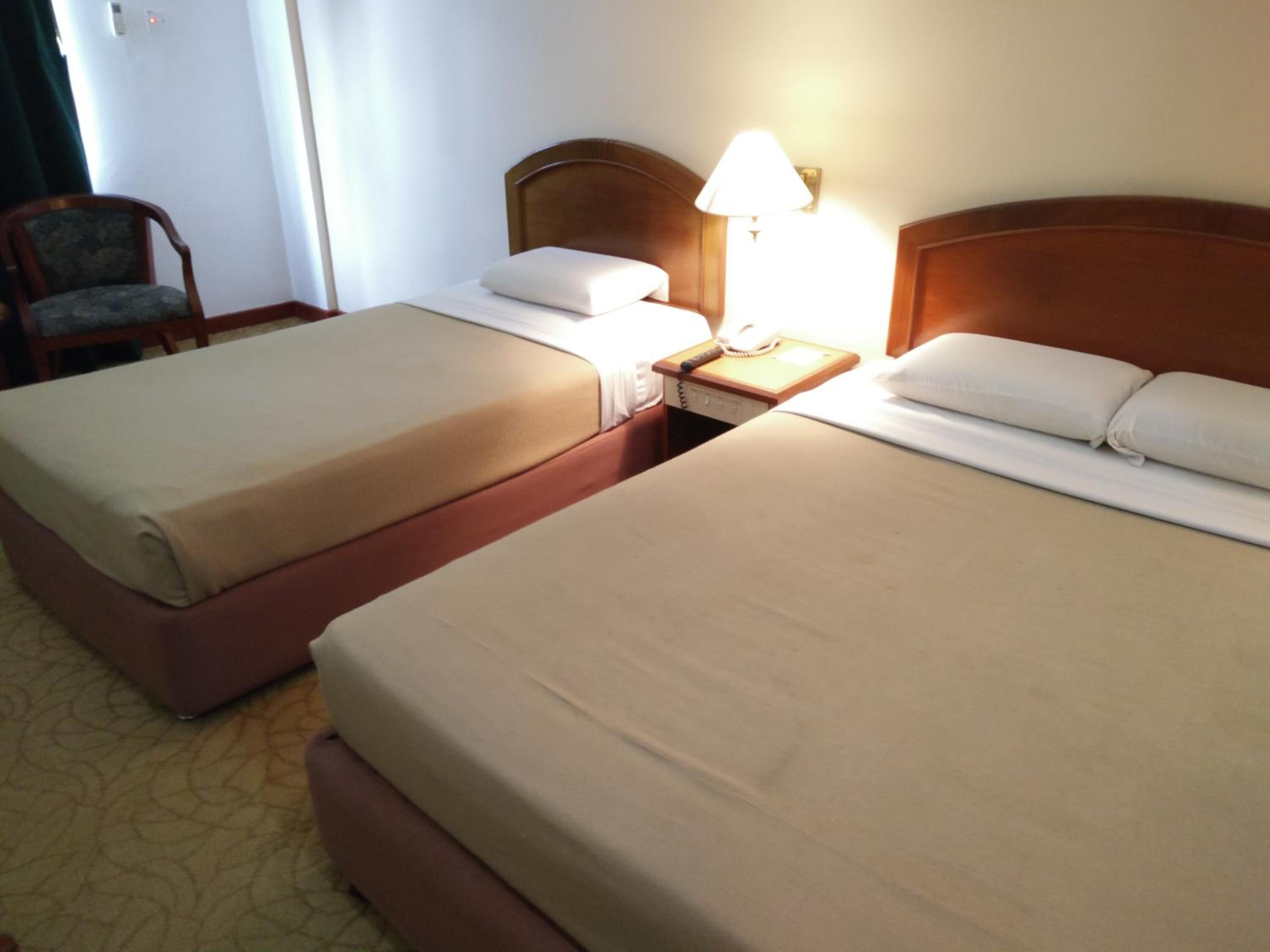 Dos camas de hotel juntas, con una mesita de noche entre ellas, una lámpara sobre la mesita y una silla al fondo.