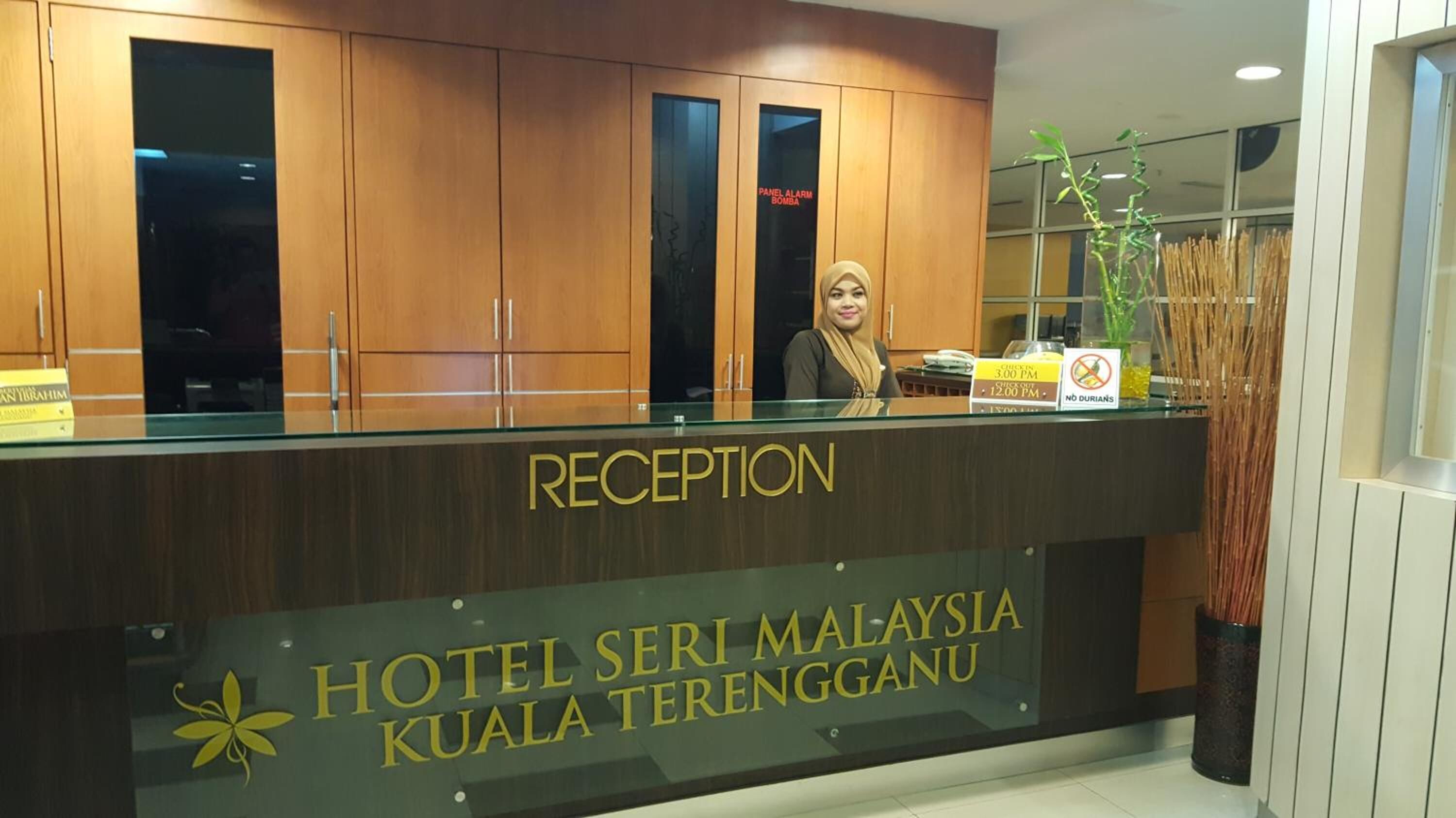Mostrador de recepción en el Hotel Seri Malaysia Kuala Terengganu, con un letrero que indica un registro de salida a las 15:00 y un registro de entrada a las 12:00.