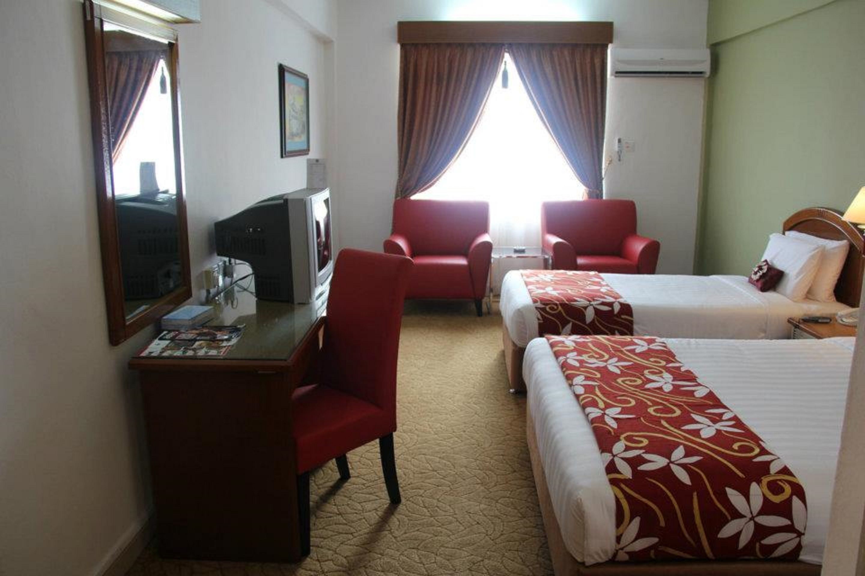 A hotel room with two single beds, a desk with a chair, a TV, and two red armchairs.