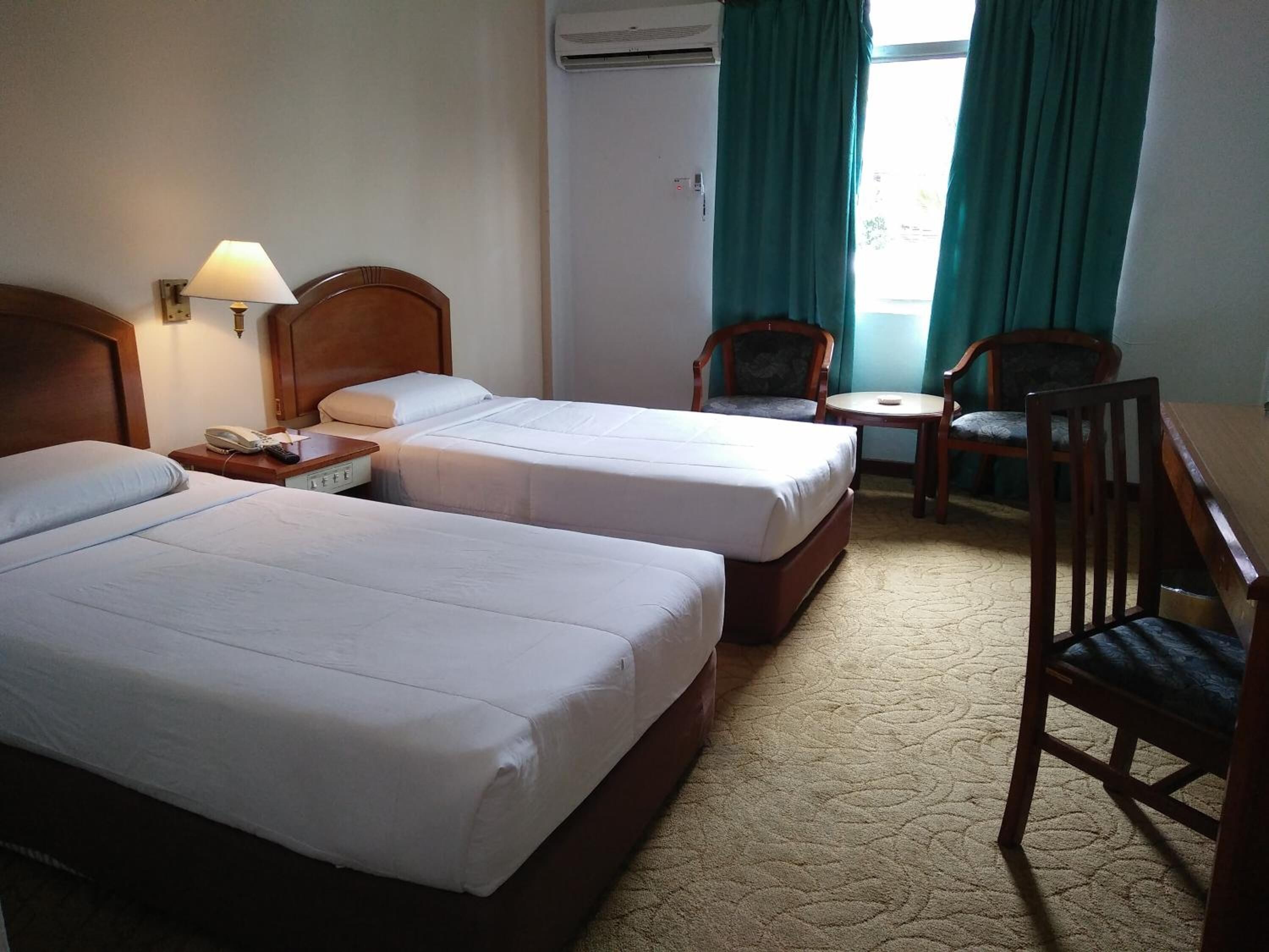 A hotel room with two beds, a desk, a chair, a lamp, and an air conditioning unit.