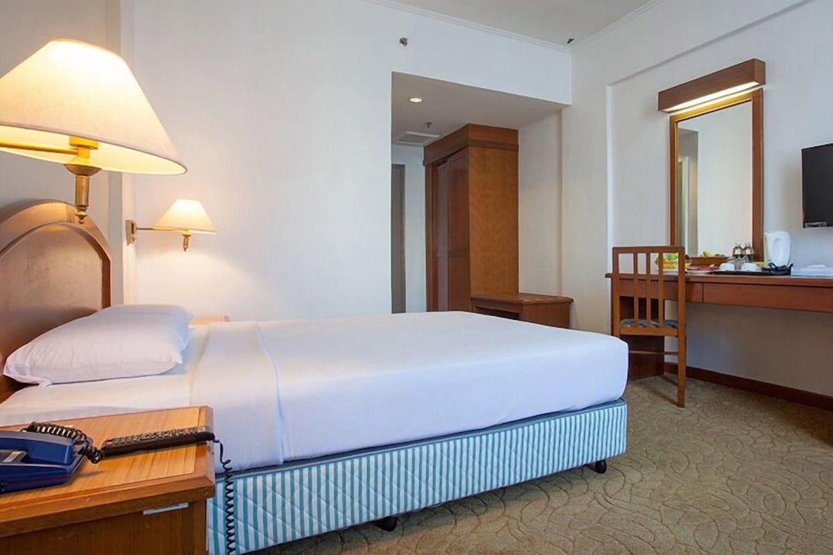 Quarto de hotel com cama, luminárias de cabeceira, escrivaninha com espelho, televisão e telefone.