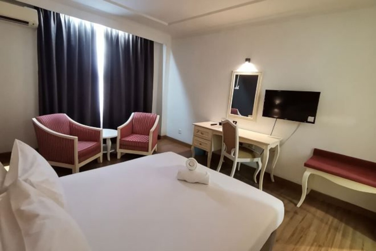 A hotel room with a bed, two red chairs, a desk, a TV, and a window with curtains.