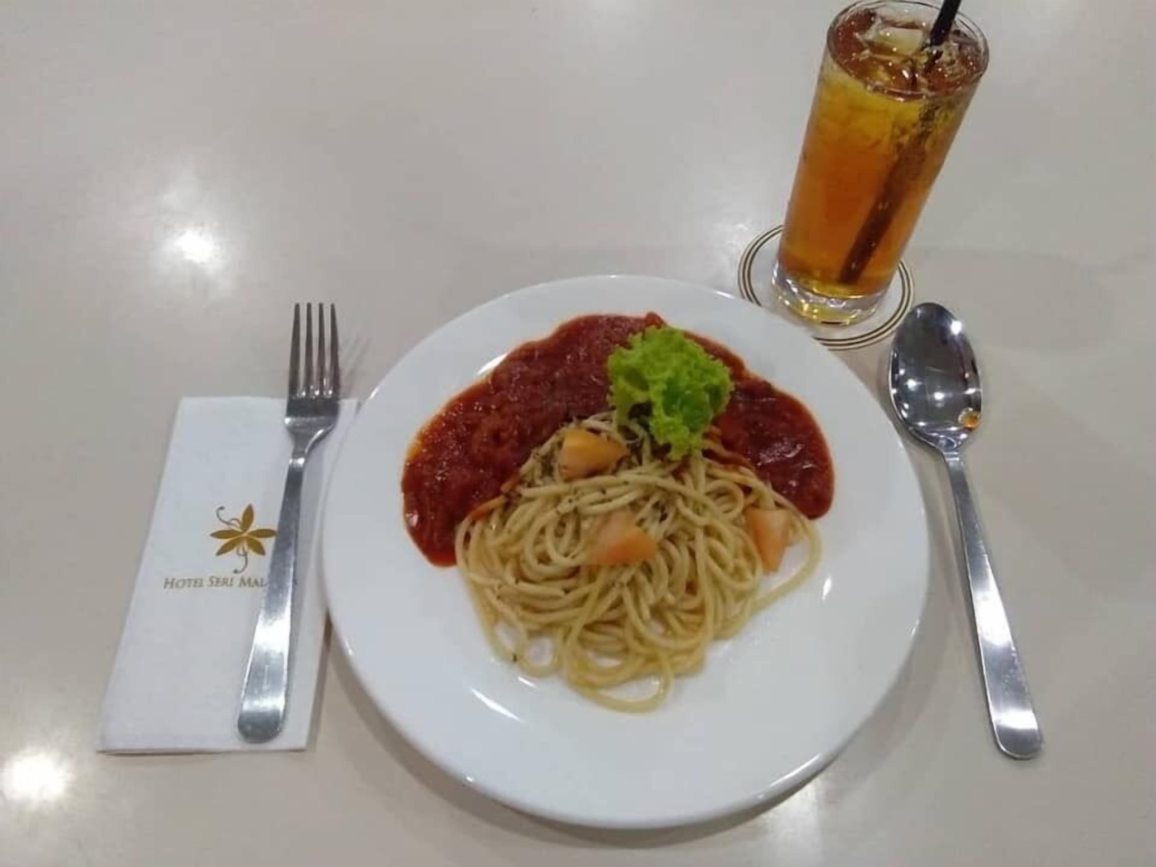 Une assiette de spaghettis à la sauce tomate, garnie d’herbes vertes, accompagnée d’un verre de thé glacé et de couverts sur une table.