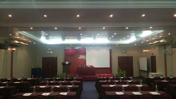 Un salón de conferencias con filas de mesas y sillas, un escenario con un atril y una pantalla al frente.