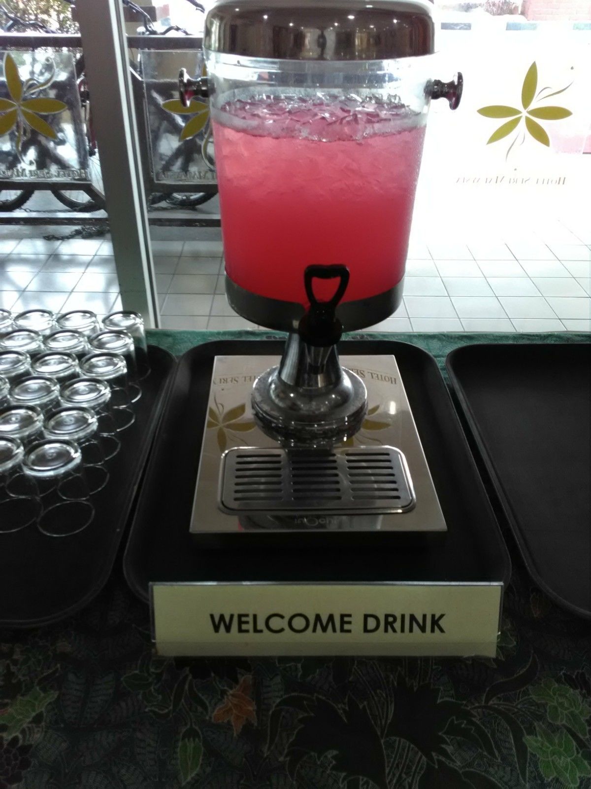 Un dispensador de bebidas con un líquido rosado, un letrero que dice 'Welcome Drink' y una bandeja con latas de bebida.