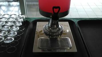 Un dispensador de bebidas con un líquido rosado, un letrero que dice 'Welcome Drink' y una bandeja con latas de bebida.